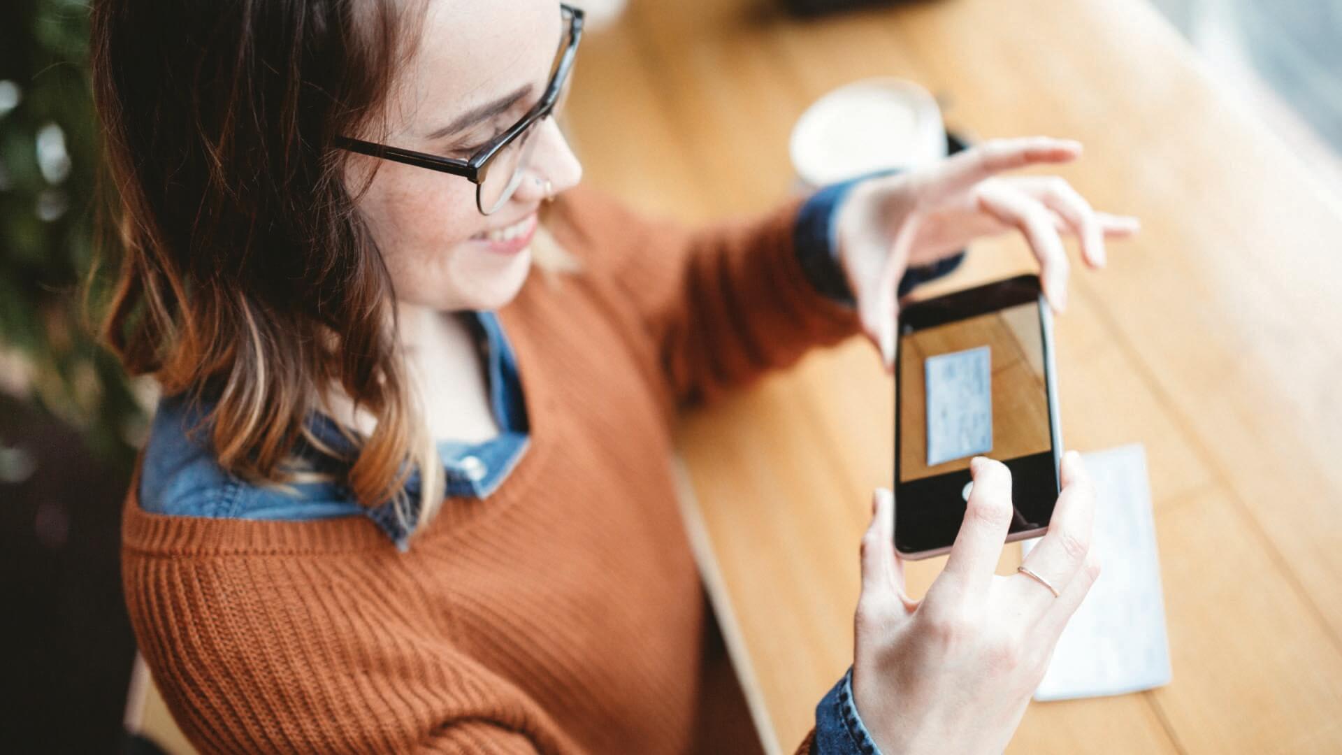 Woman sitting at table, using her phone to take a picture of a check and deposit it remotely.