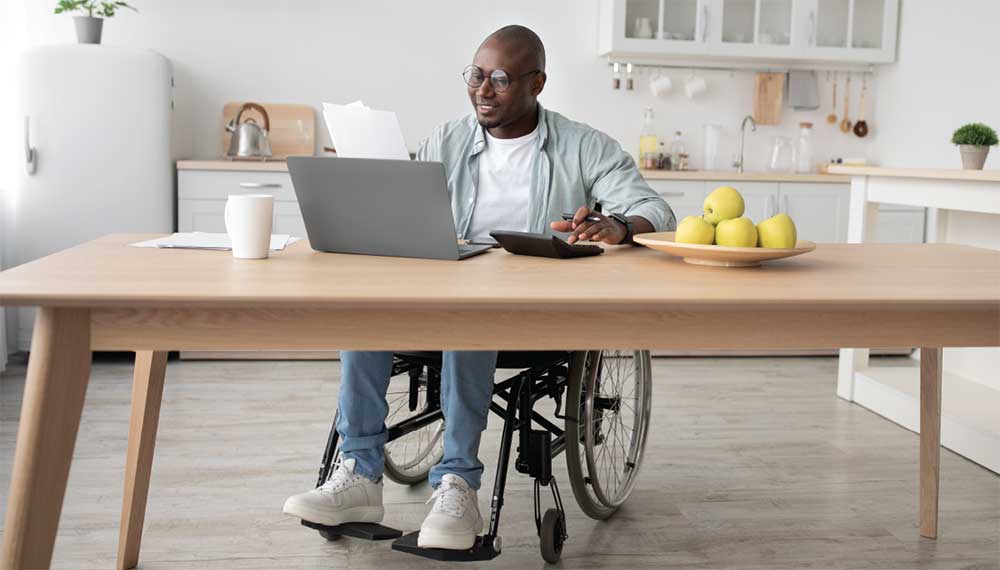 African American man sits in a wheel chair at a table in front of a laptop.