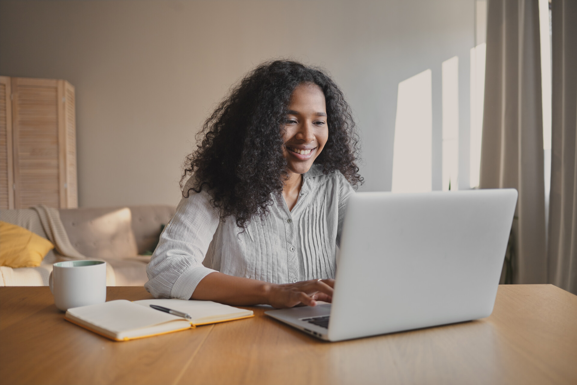 Cheerful young African American woman copywriter sitting in front of open laptop with mug and copybook on desk, checking estatements.
