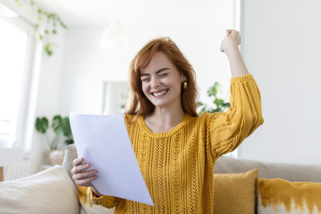 Excited, smiling young woman holds a paper with her right hand while making an excited fist motion with her left hand while sitting on a couch.