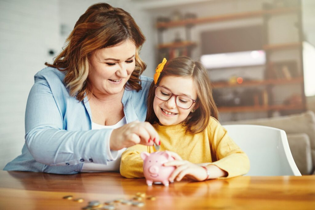 A woman and a girl happily adding coins to a piggy bank, illustrating a fun way to teach kids about saving money.