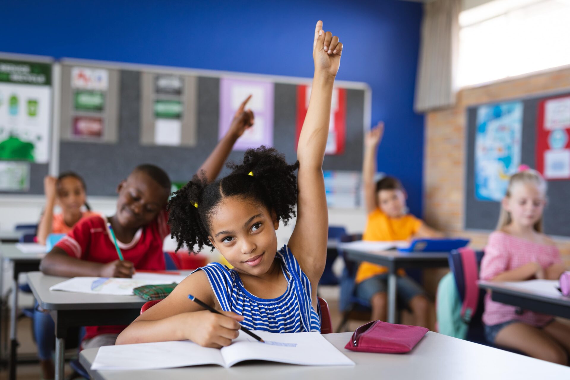 A young girl eagerly raises her hand in class, ready to answer a question and turn in her report card for report card, a benefit of having a youth account at Park View.