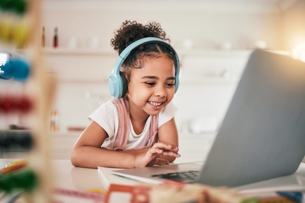 A cheerful girl wearing headphones uses a laptop, happily exploring online while staying safe from potential scams.
