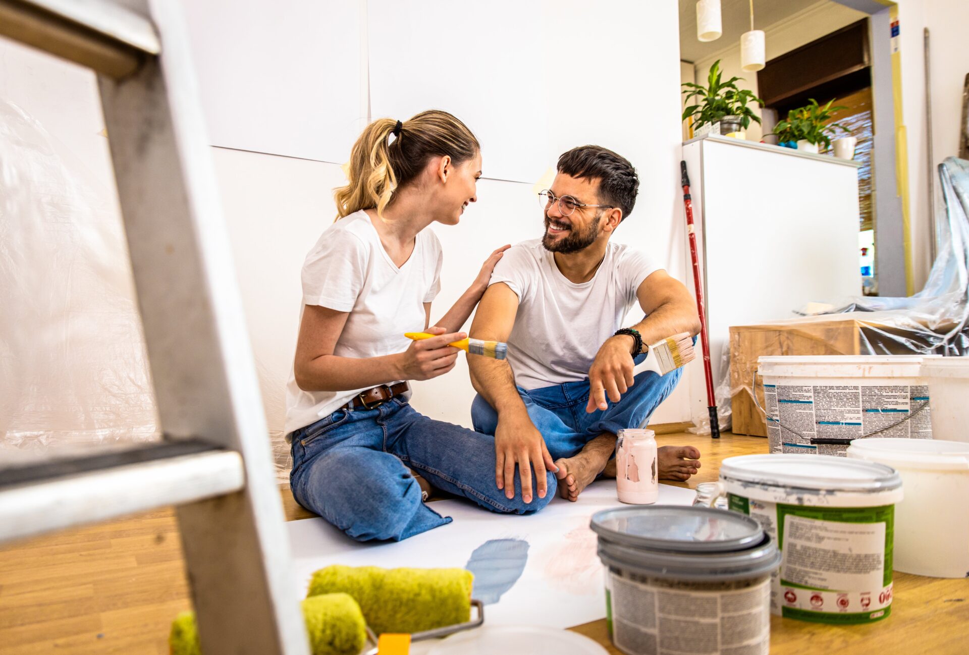 A couple smiles at each other while holding paint brushes surrounded by paint cans, using the equity in their home to do a home renovation.