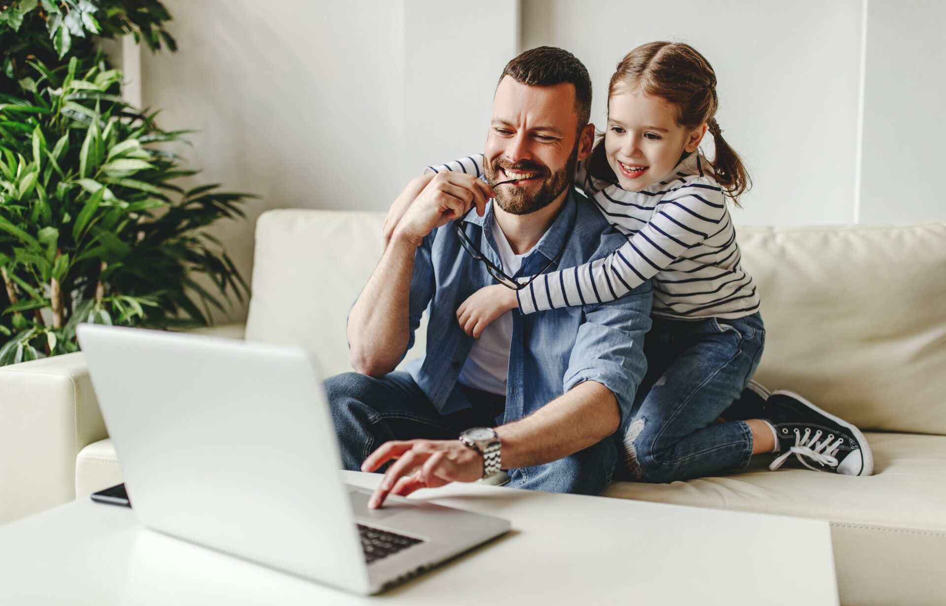 Father and daughter sit on a sofa, smiling as they look at a laptop. The girl leans on her dad's shoulder, creating a warm and joyful atmosphere.
