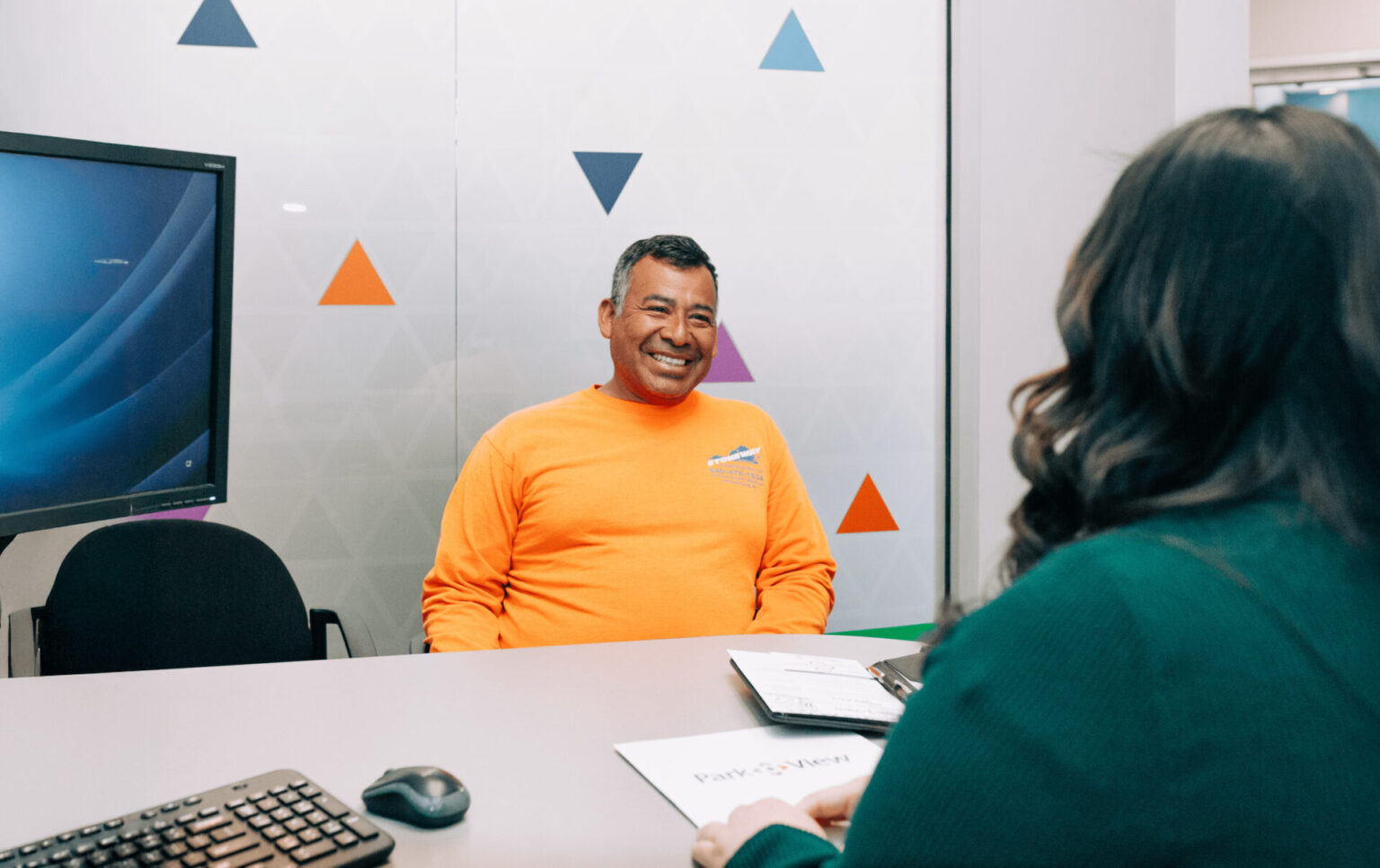 Smiling, middle-aged man in orange shirt sits at desk across from a Park View employee opening a Park View checking account.