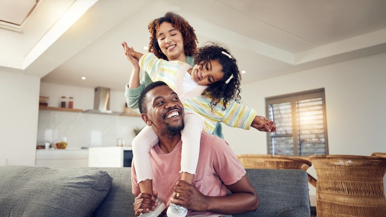 Family, father and daughter playing with mother on living room sofa together for fun bonding at home. Happy dad carrying child for piggyback ride with mom on lounge couch.