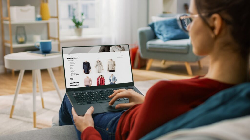 Young Woman at Home Using Laptop Computer for Browsing Through Online Retail Shopping Site. She's Sitting On a Couch in His Cozy Living Room. Over the Shoulder Camera Shot