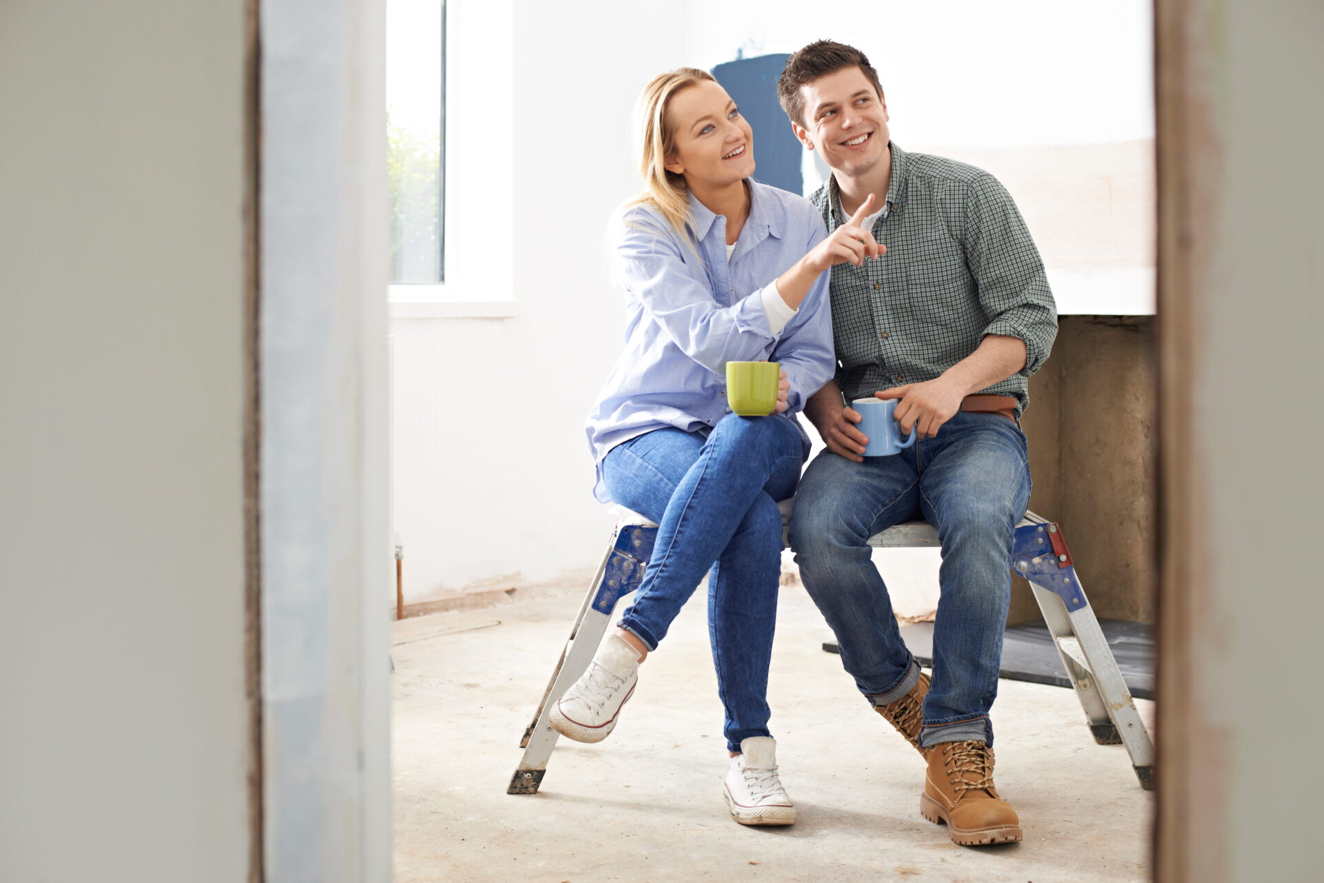 A woman and a man sit on a ladder in a partially renovated room, discussing their plans. The woman holds a mug and gestures as they smile, envisioning how their home equity can help them complete the remodel.