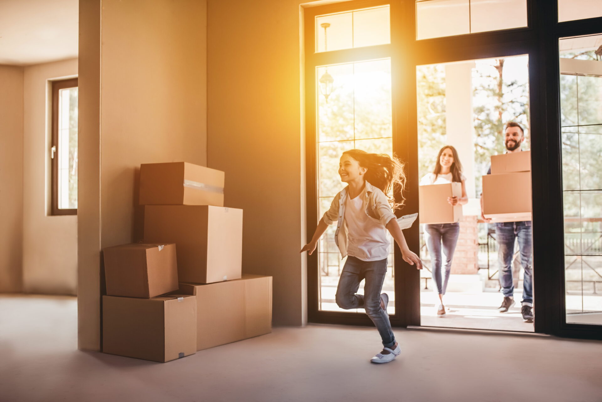 A young girl joyfully runs inside a sunlit room with cardboard boxes piled on the left. A smiling couple enters, carrying more boxes, conveying excitement.