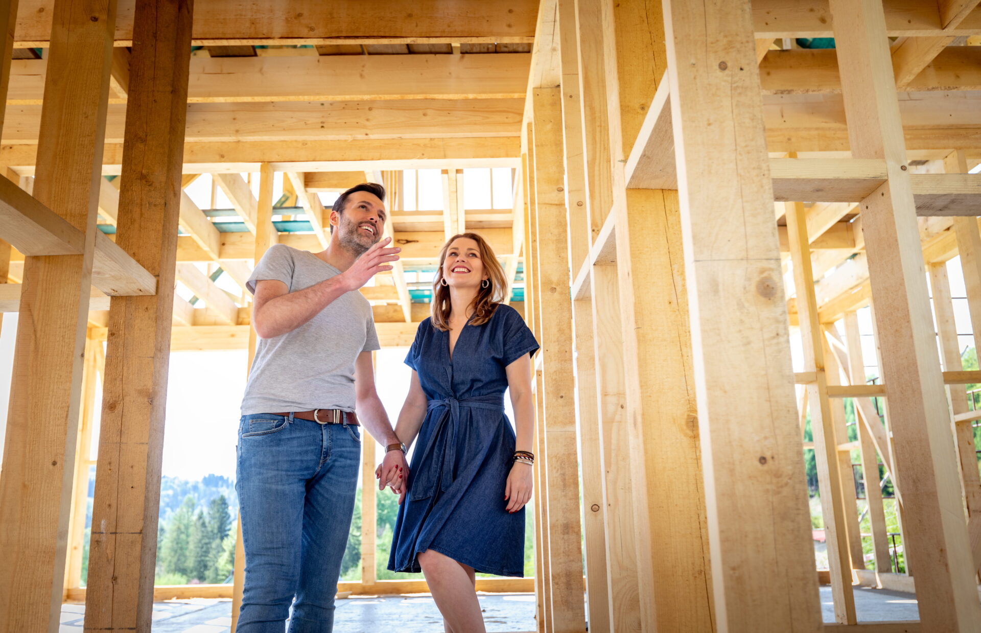 A man and woman stand inside their wooden house under construction, holding hands and smiling, as sunlight streams through the unfinished framework—celebrating a new chapter made possible by their construction loan.