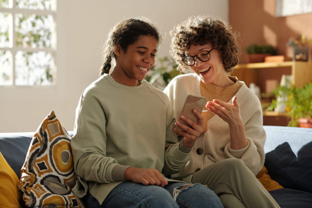 Happy mom spending time with her adopted daughter at home, they sitting on sofa and watching photos on mobile phone
