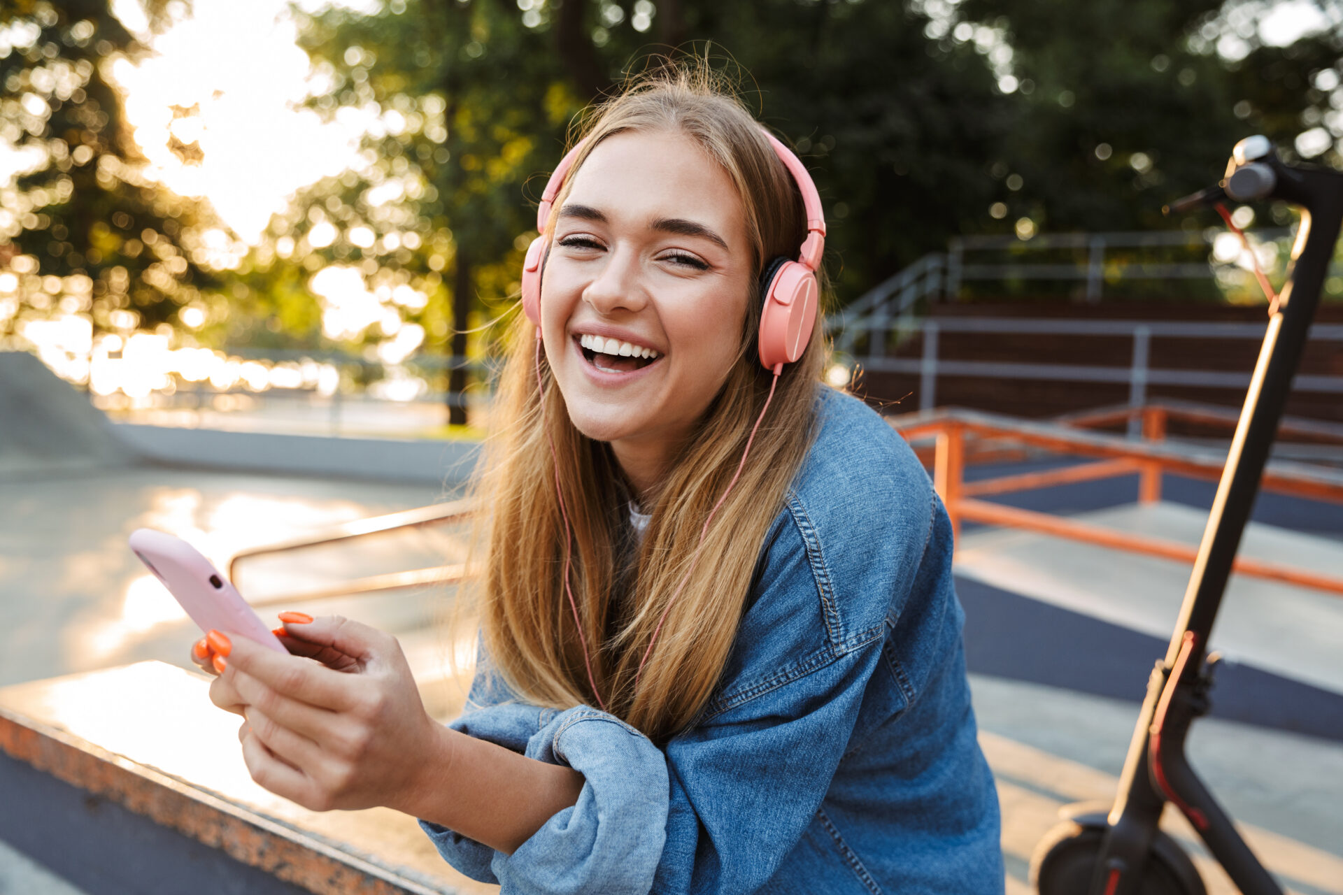 A smiling young woman wearing pink headphones sits outdoors at a skate park, holding a smartphone. She’s dressed casually in a denim jacket, with a scooter beside her and trees in the background, enjoying music in warm sunlight.