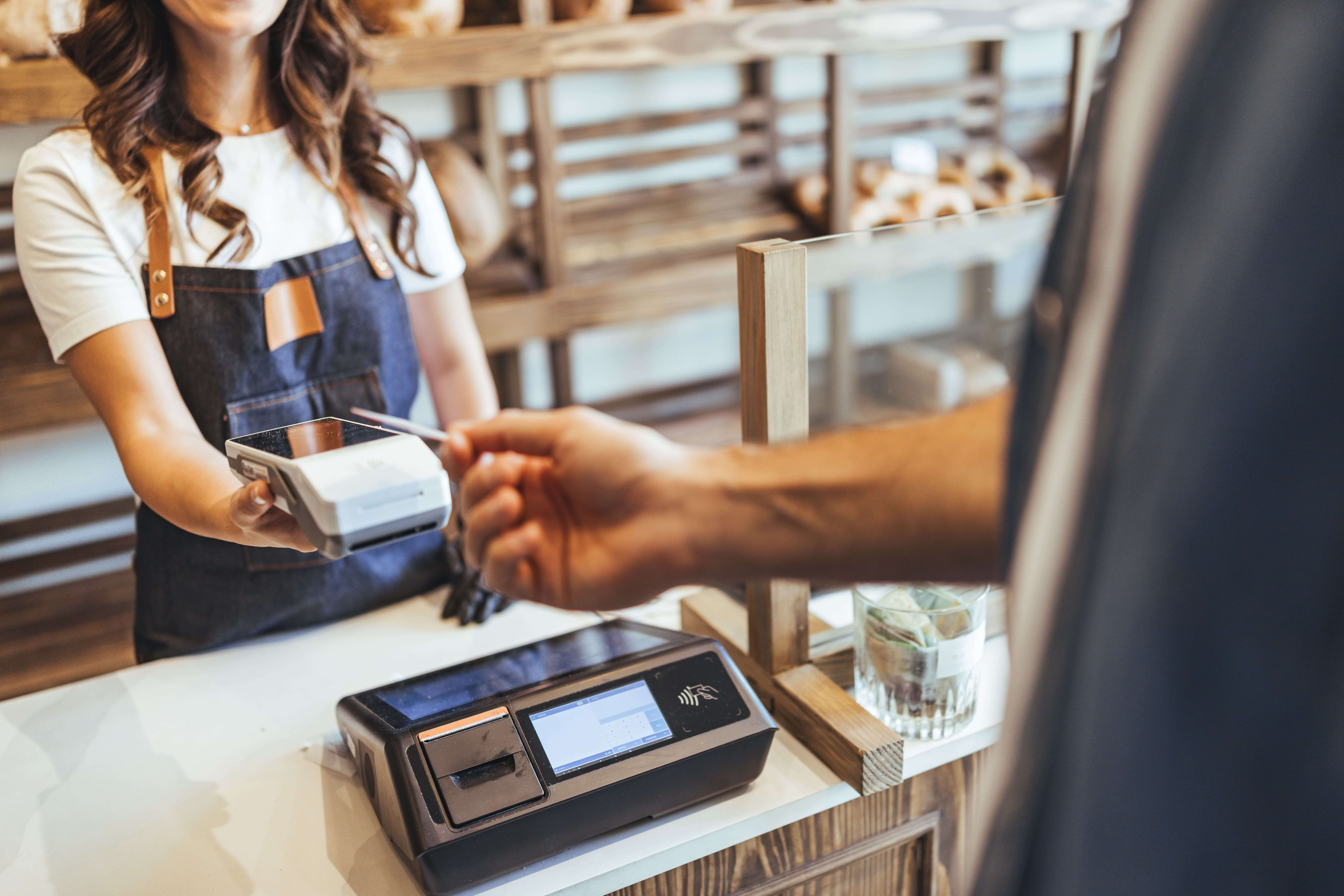 A cashier in a bakery hands a card reader to a customer. The setting is warm, with wooden shelves and baked goods in the background. Payment in progress.