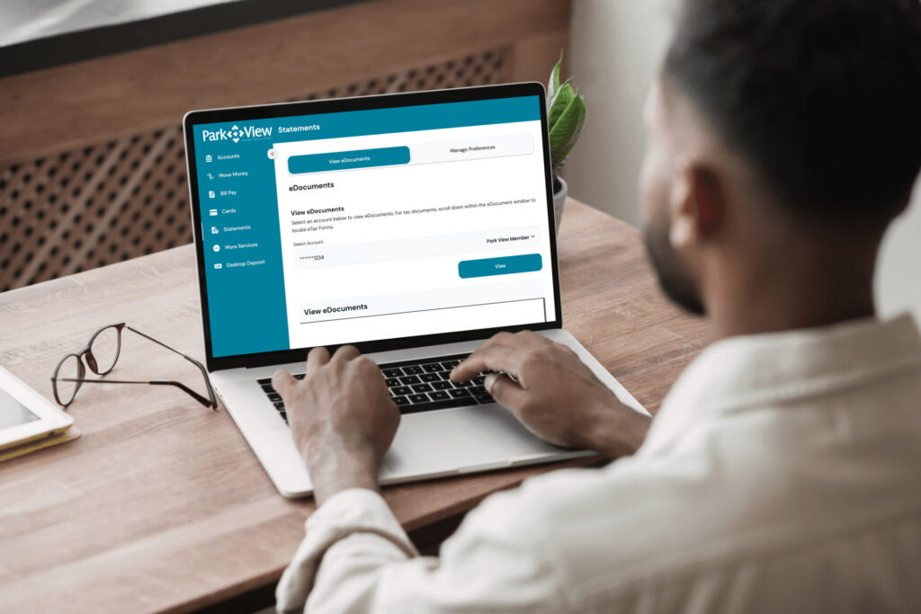 A man sits at a wooden table using a laptop displaying a financial management webpage with tabs like "eDocuments." Glasses lie beside the laptop.