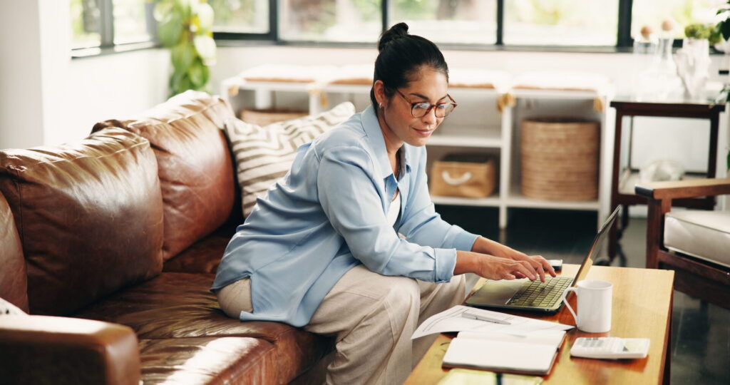A woman sits on a couch, focused on her laptop, reviewing her mid-year financial check-in.