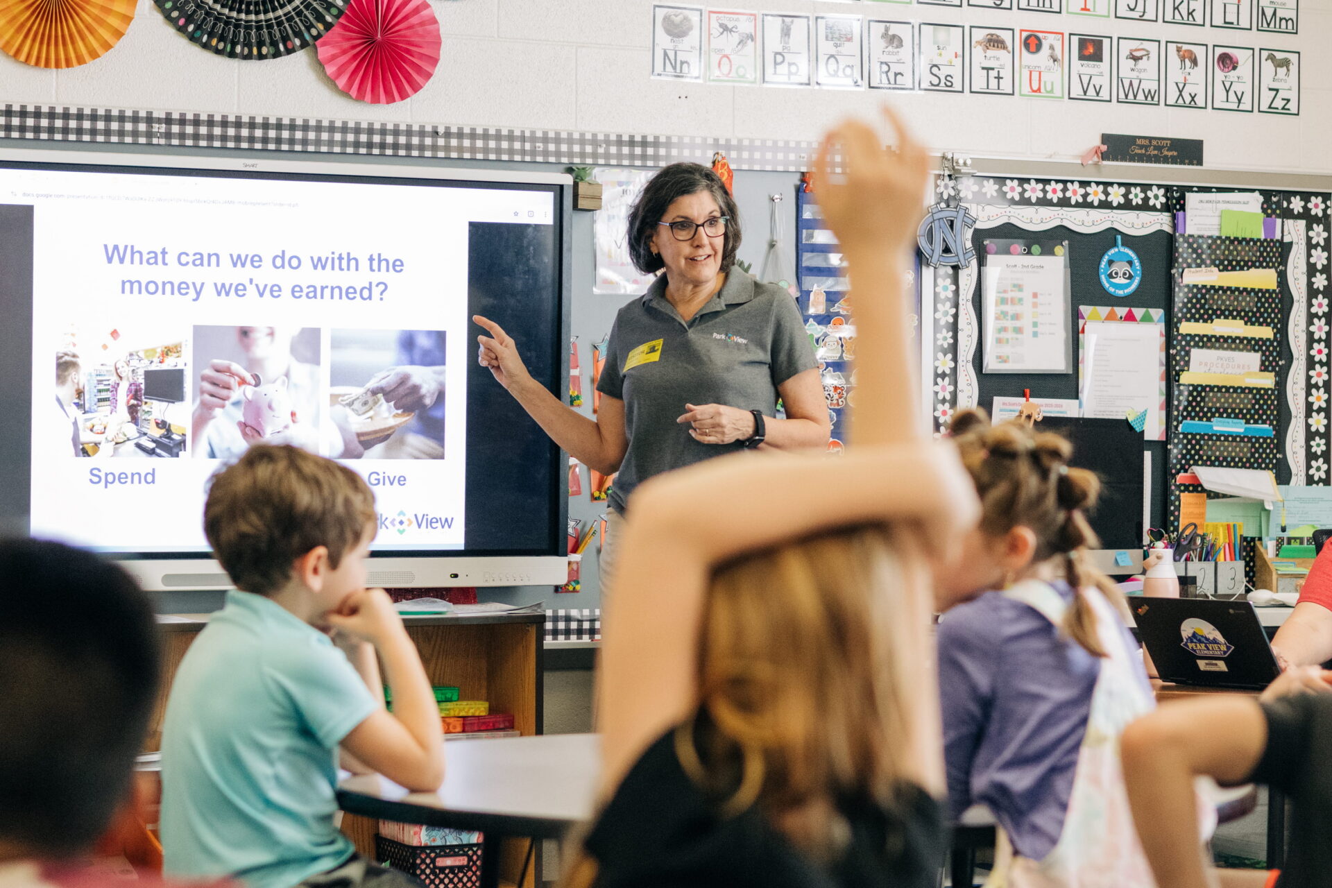 A woman in a gray shirt points to a board in a school classroom, sharing about financial education from Park View Federal Credit Union.