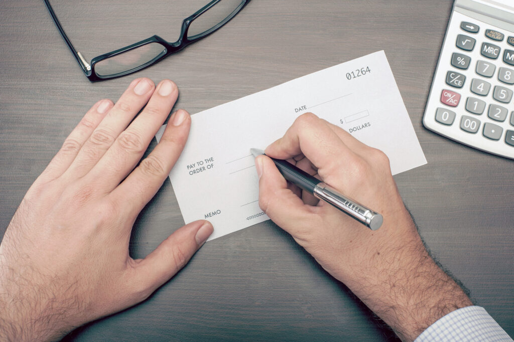 Top-down view of hands filling out a financial check with a pen on a desk, beside eyeglasses and a calculator; conveys focused money management.