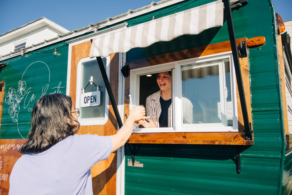 A smiling vendor serves a customer through the open window of a green food truck; sunlight and a striped awning create a warm, welcoming street scene.