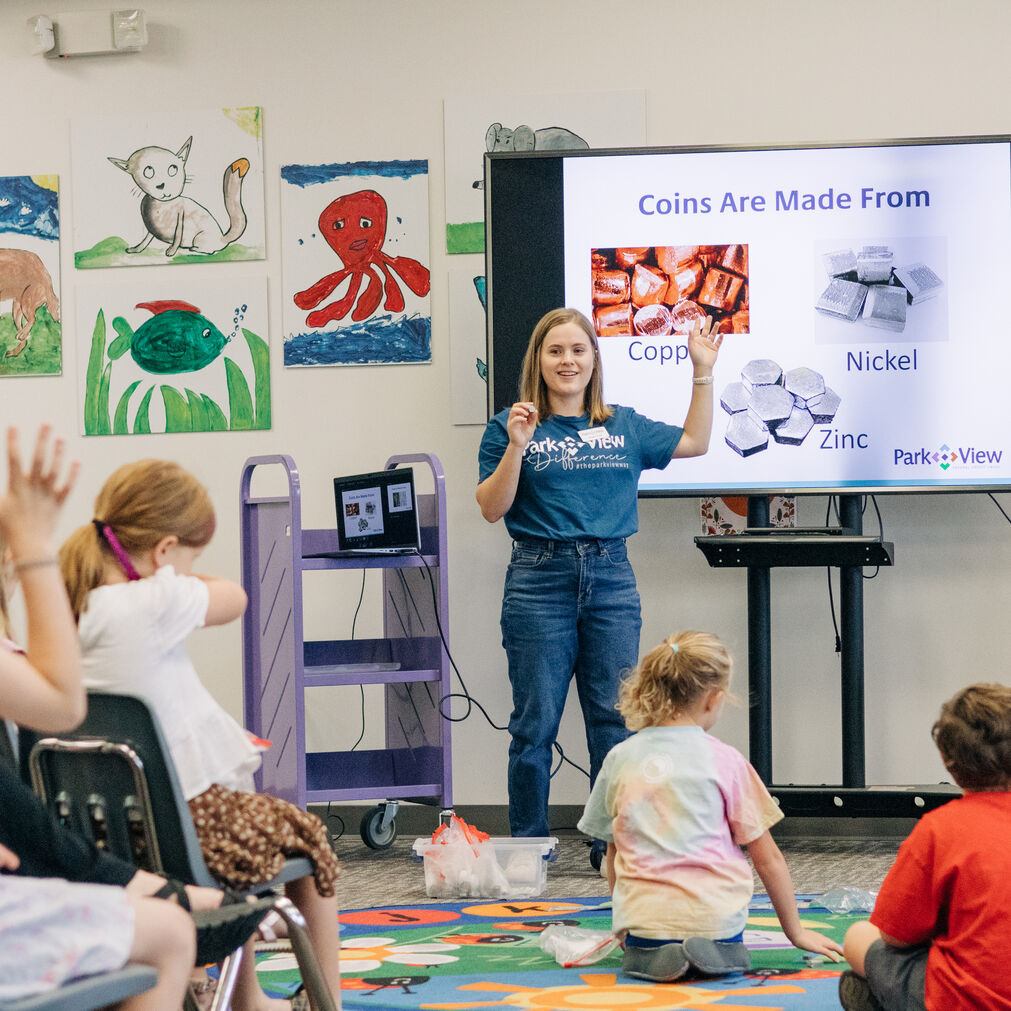 Young presenter explains how coins are made from copper, nickel, and zinc to children in a colorful, engaging classroom activity.