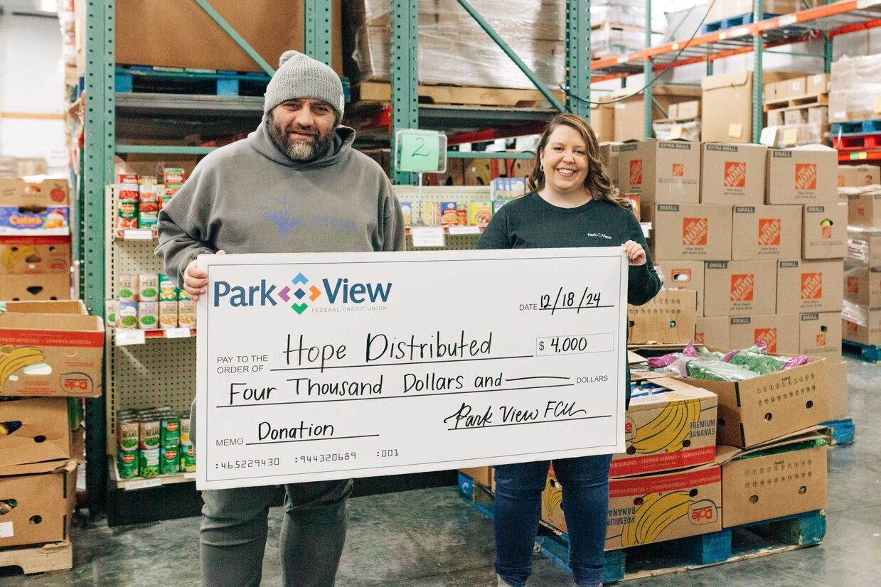 Two smiling people hold a $4,000 Park View donation check for Hope Distributed in a busy warehouse, conveying generosity and community support.