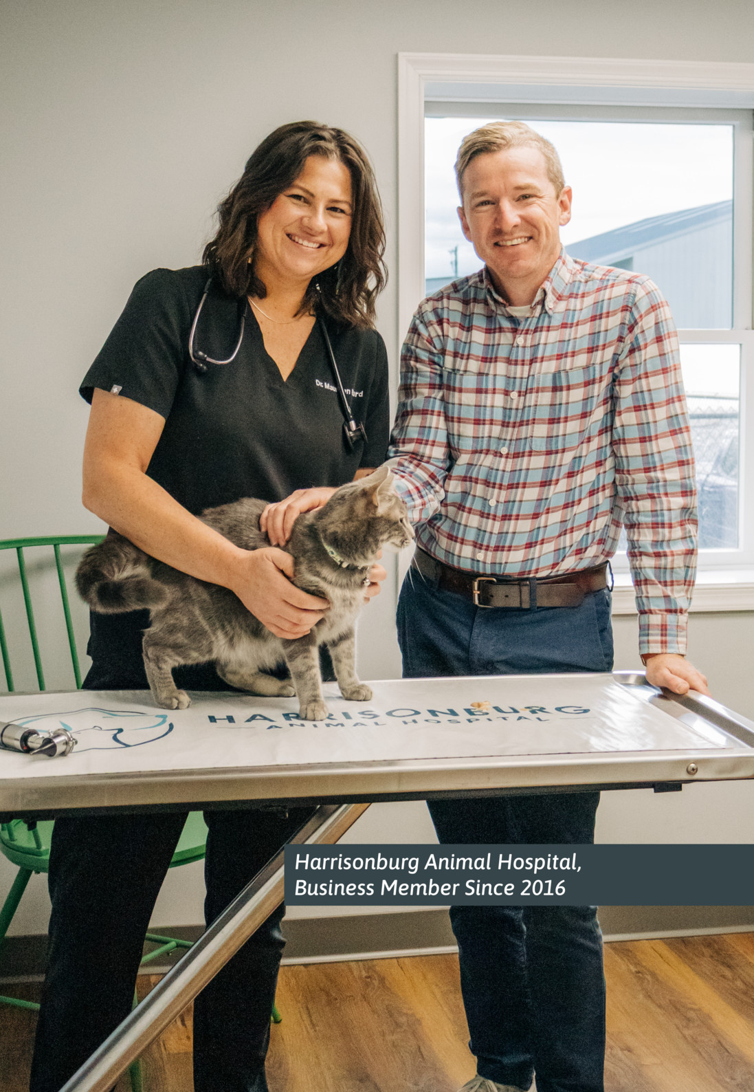 A smiling veterinarian and man pose with a gray cat on an exam table at Harrisonburg Animal Hospital, conveying a warm, caring atmosphere.