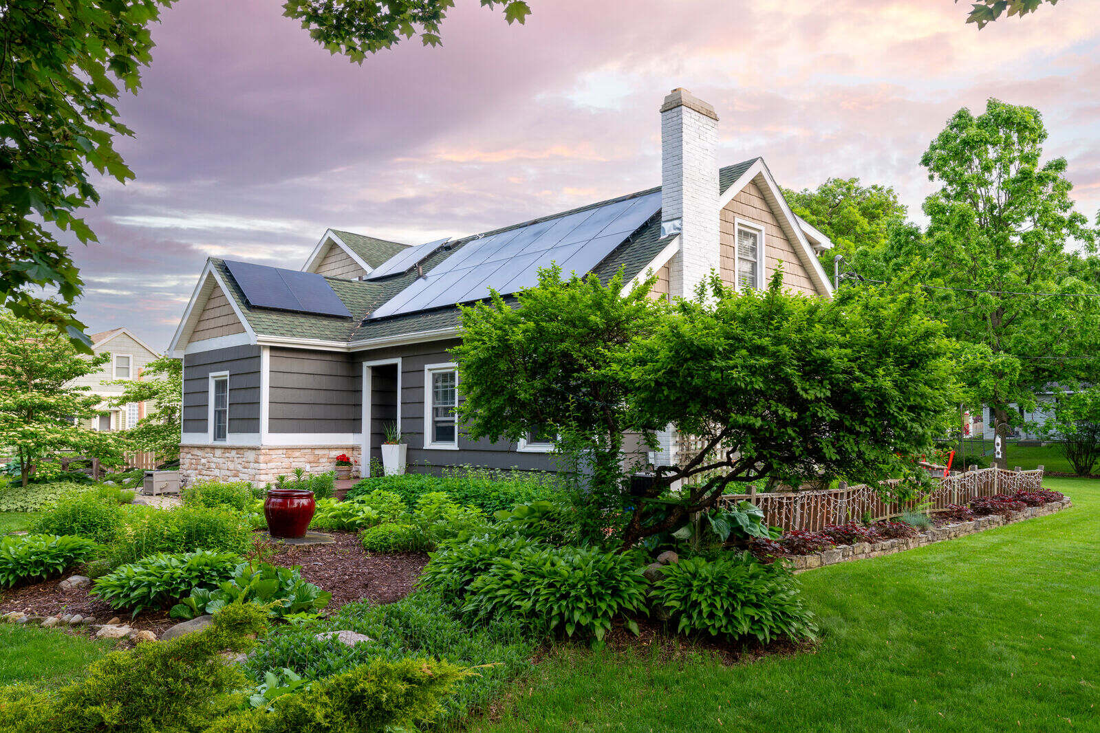 Gray suburban home with solar-panel roof, lush gardens, trimmed lawn, and leafy trees beneath a softly colored evening sky.