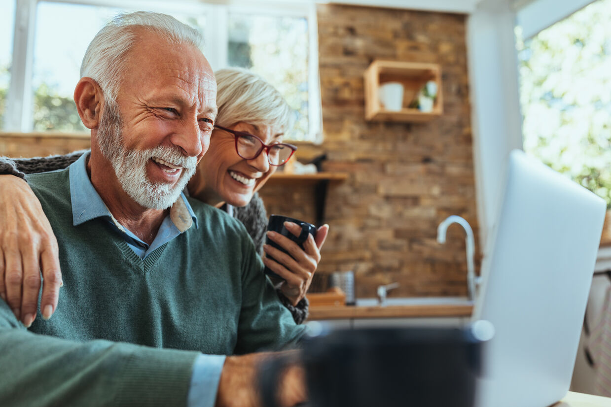 Smiling older couple sharing a joyful moment while looking at a laptop in a bright, cozy kitchen, conveying connection and happiness.