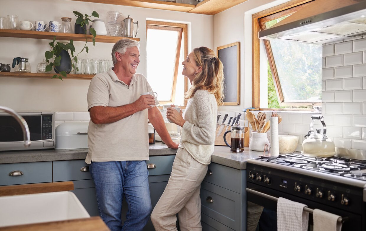 Smiling older couple chat together in a bright, stylish kitchen, holding mugs near open shelves and a stove, creating a warm, relaxed mood.