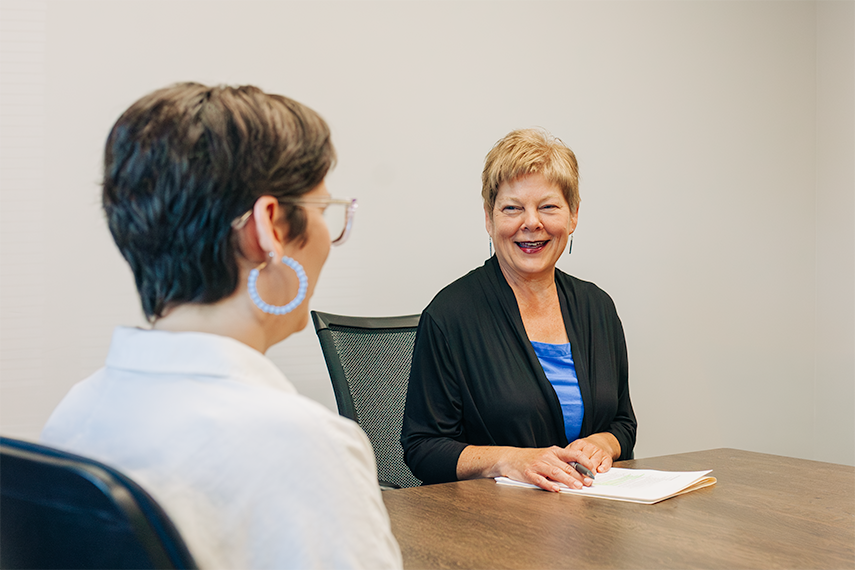 Two women sit at a table in an office setting, facing each other. Park View financial advisor Debbi Watcher holds papers and smiles as she listens to the other, suggesting a professional meeting.