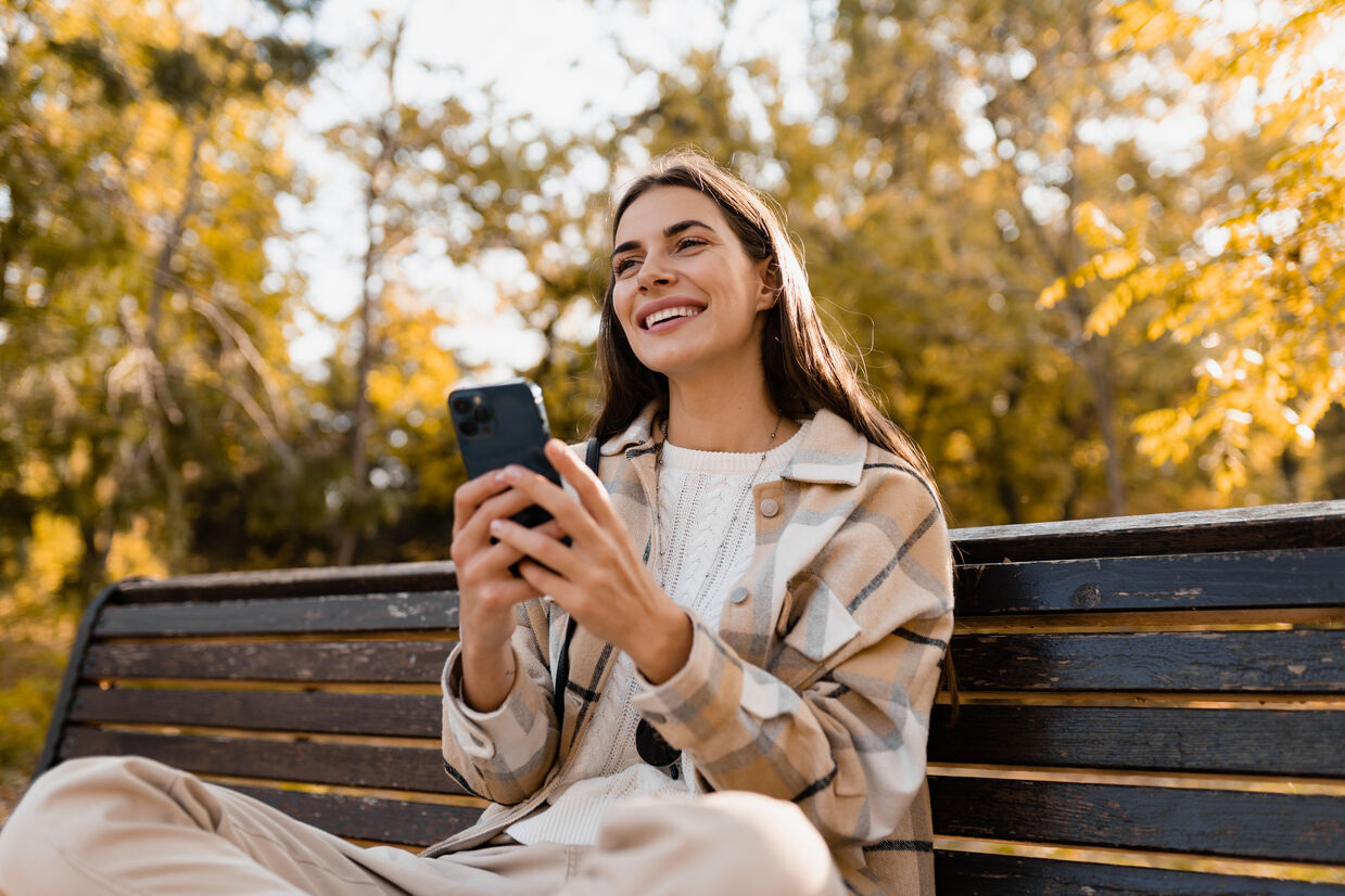 Smiling young woman in a plaid jacket uses a smartphone on a park bench, surrounded by golden autumn trees, conveying warmth and joy.