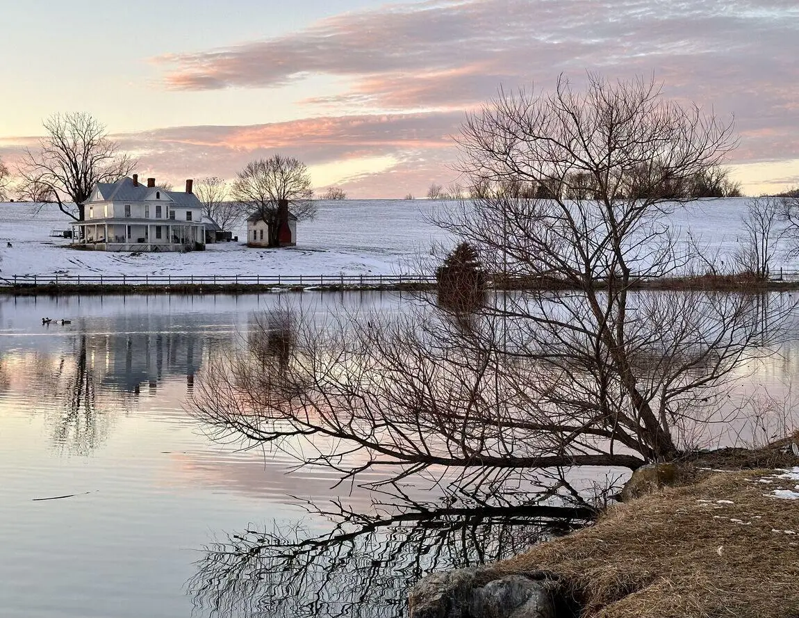 A cozy house by a snowy lake, surrounded by white snow and calm water, creating a peaceful winter scene.
