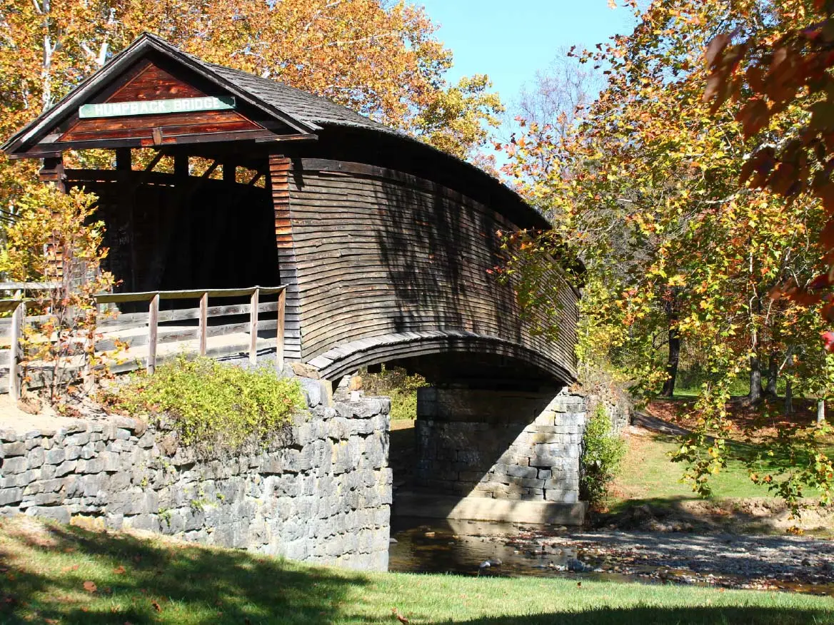 A rustic wooden covered bridge spans a serene river, surrounded by lush greenery and a clear blue sky in Covington, VA