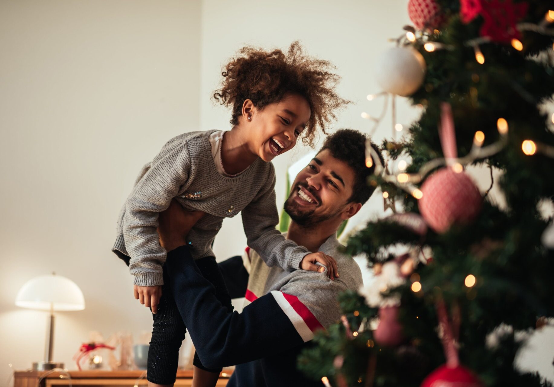 A man lifts a laughing child in front of a beautifully decorated Christmas tree, capturing the joy and warmth of the holiday season.