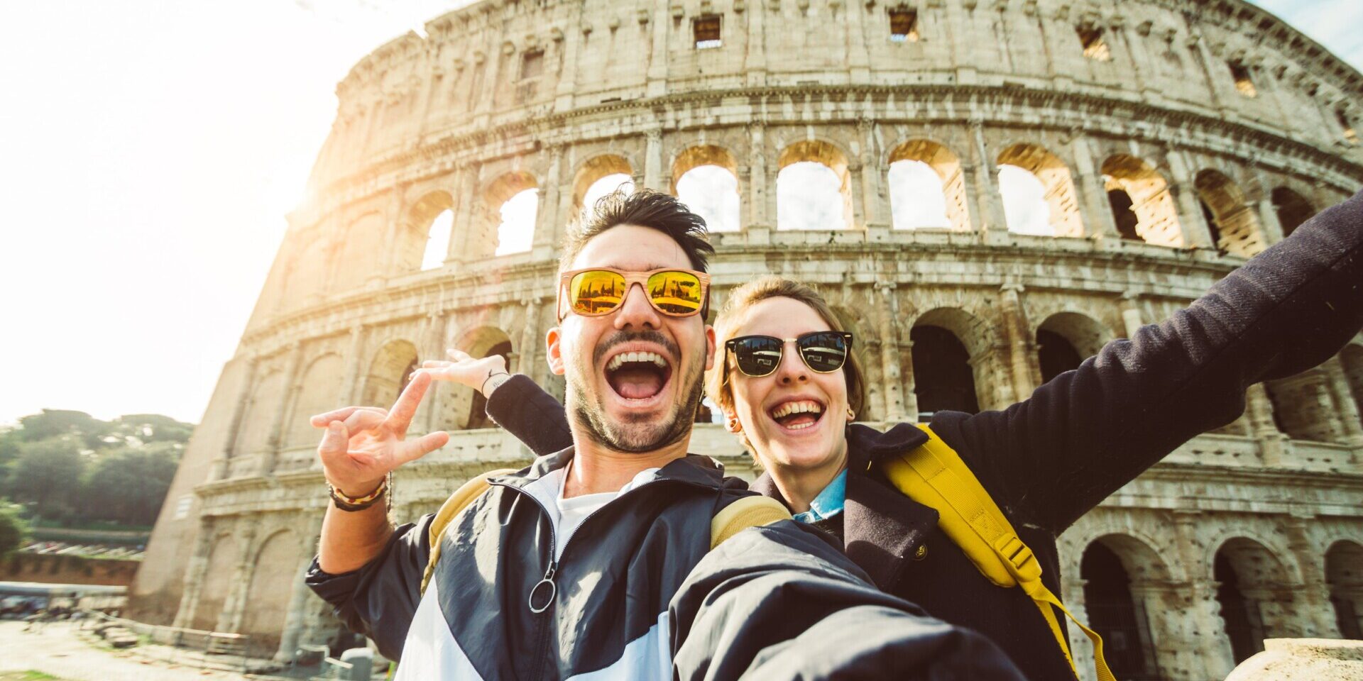 Two tourists smiling and taking a selfie in front of the iconic Colosseum in Rome, capturing a memorable moment.