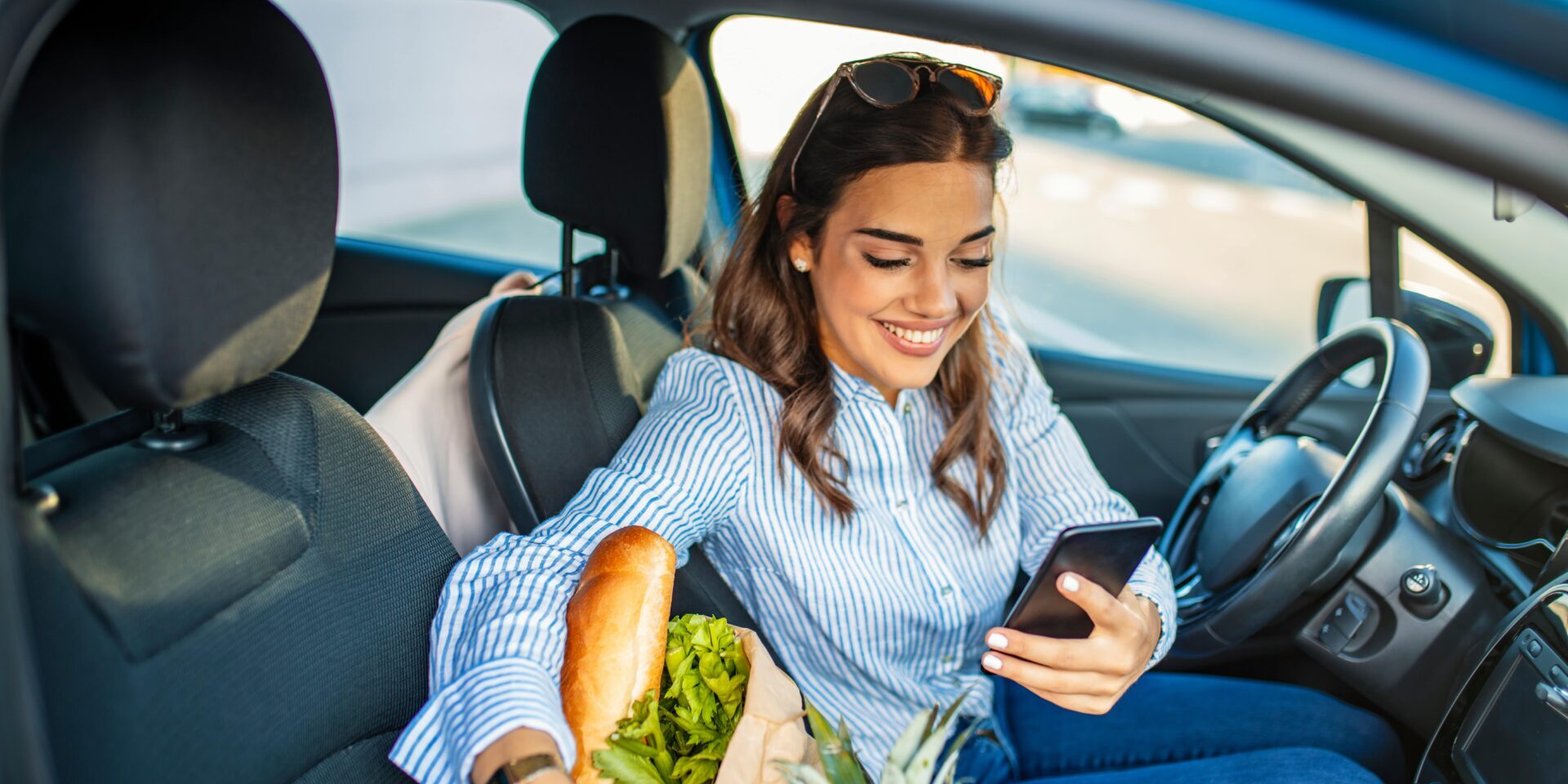 A woman sits in her car and smiles at her phone while groceries sit in the passenger seat.