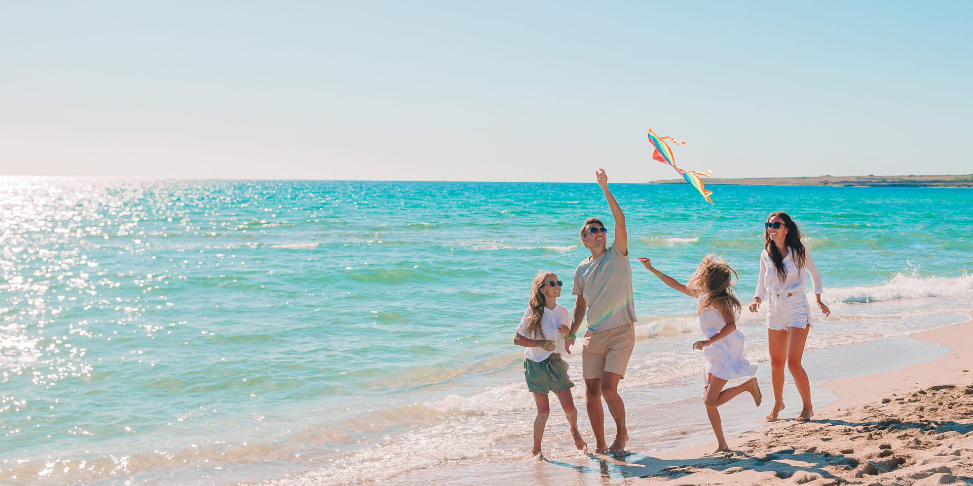 Four people enjoy a sunny beach; one adult flies a colorful kite while two children run nearby, and another adult strolls beside them—making memories that could be yours with a vacation loan. The ocean waves roll gently in the background.