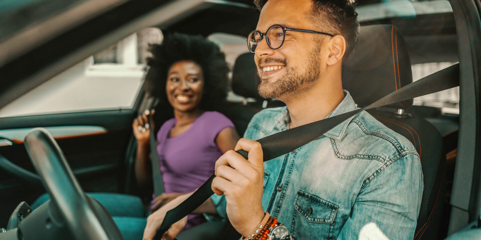 A man and woman sit in the driver and passenger seat of a car, discussing their auto loan options.