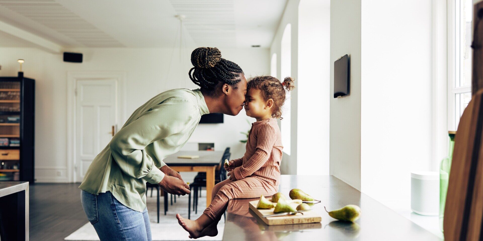 Mother and daughter share a tender forehead touch in a bright kitchen, with fresh pears on the counter