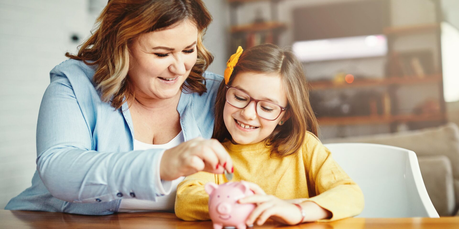 A woman and a girl happily adding coins to a piggy bank, illustrating a fun way to teach kids about saving money.