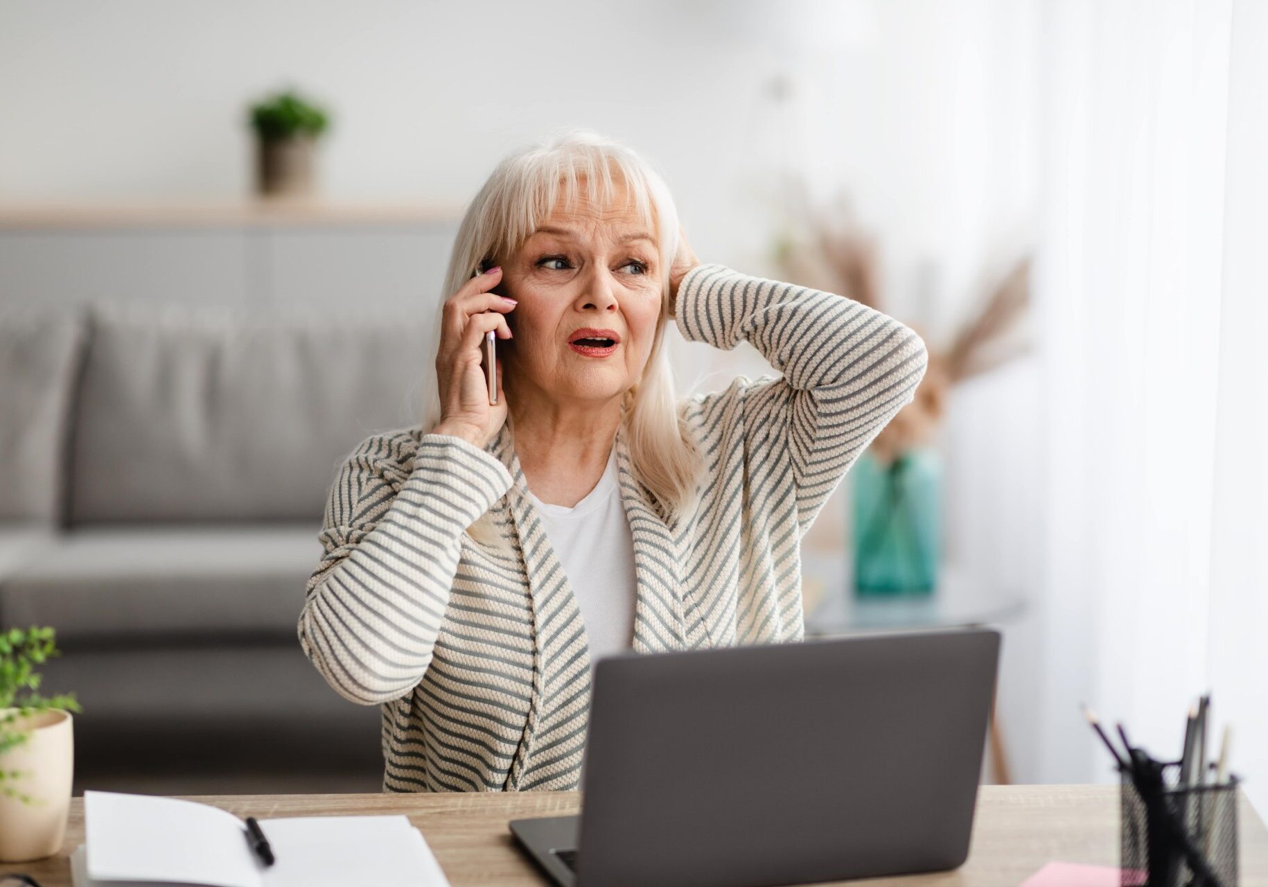 AdobeStock_477253429-min Elderly woman talking on phone at home, potential senior scam or fraud situation.