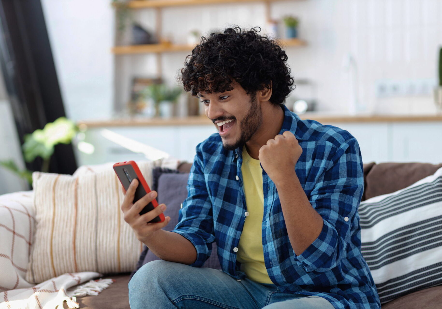 Overjoyed Indian man looking at the screen of mobile phone making yes gesture, feeling excited after receiving early direct deposit.