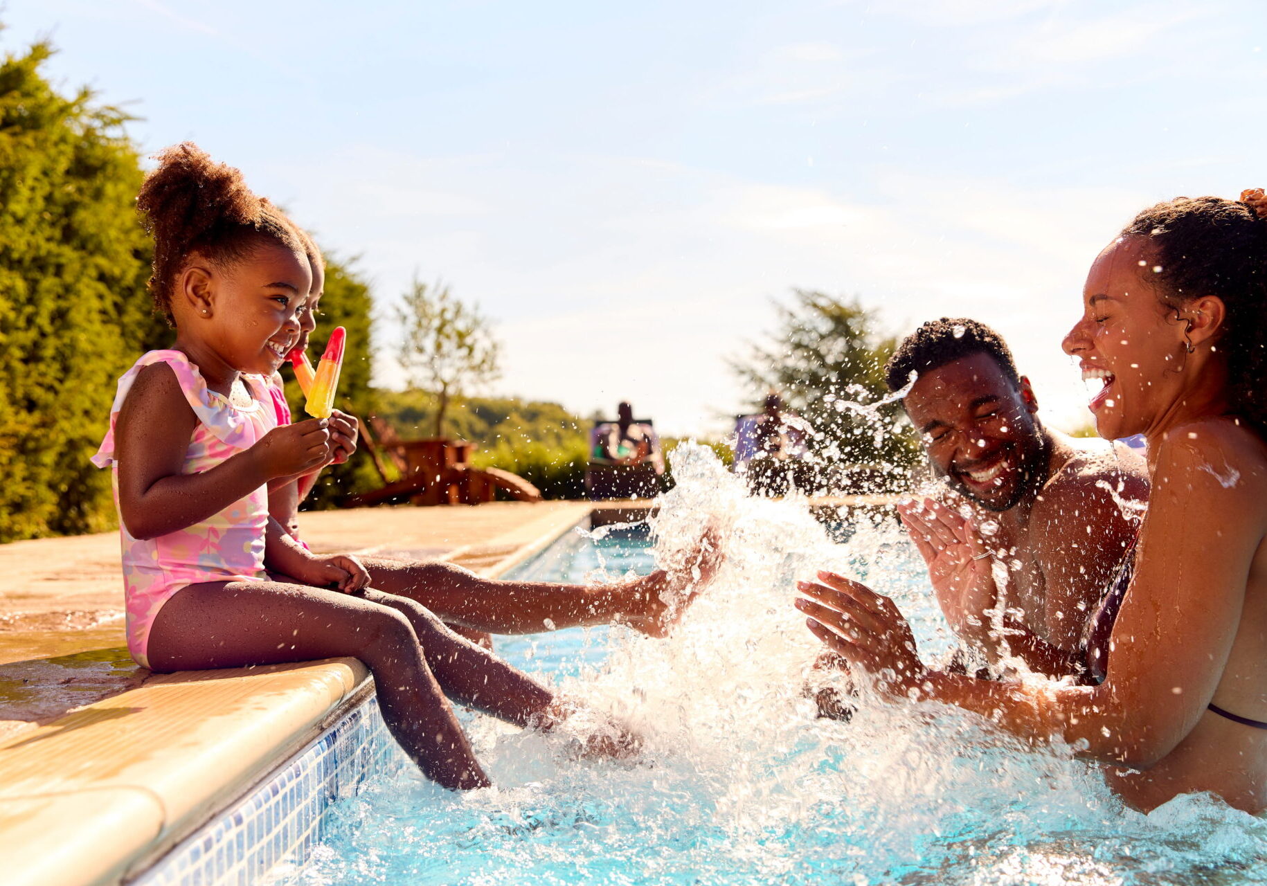 Two young children sit on the side of a pool holding a popsicle and splashing the water while a man and woman stand in front of them smiling and laughing.