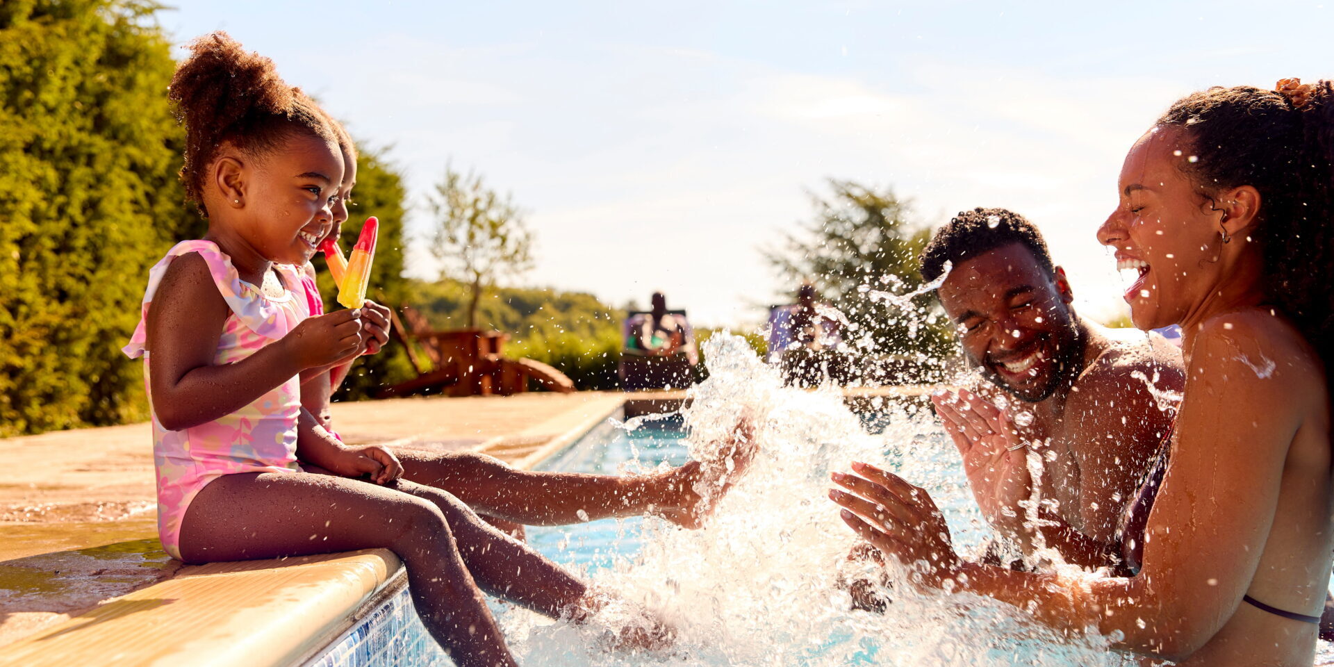 Two young children sit on the side of a pool holding a popsicle and splashing the water while a man and woman stand in front of them smiling and laughing.