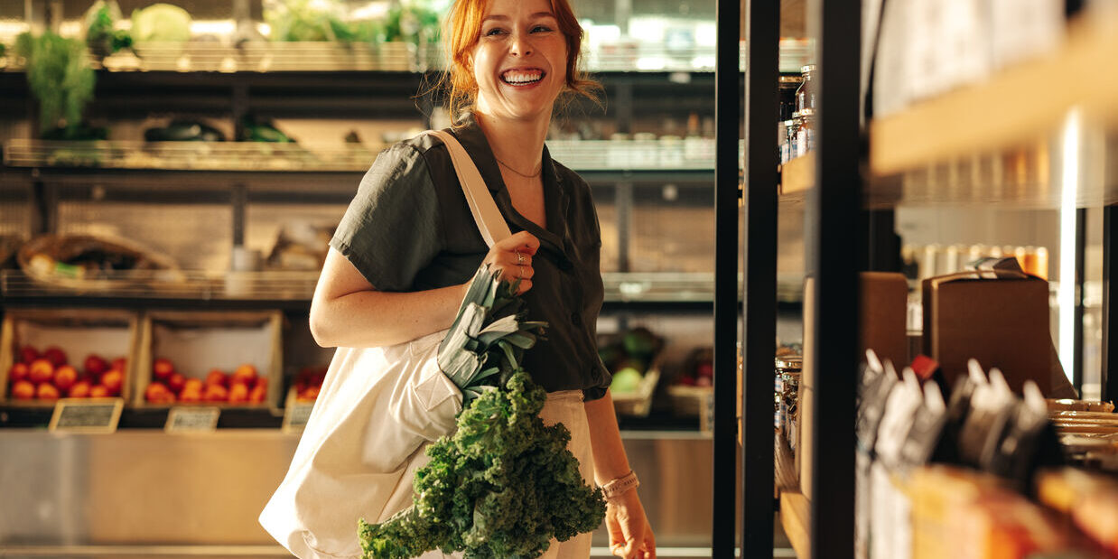 A woman stands in a grocery store, holding a bag of vibrant greens, excited about her fresh produce selection.