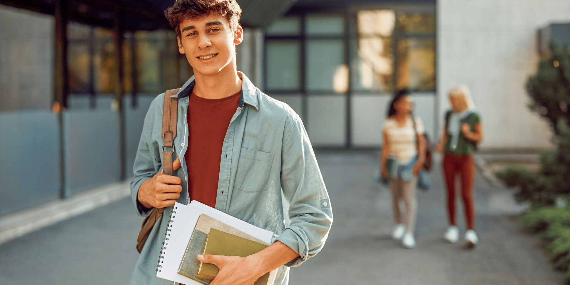 Smiling young man holds books and a backpack, standing outside a modern school building. Two people walk in the background. Bright, sunny day.