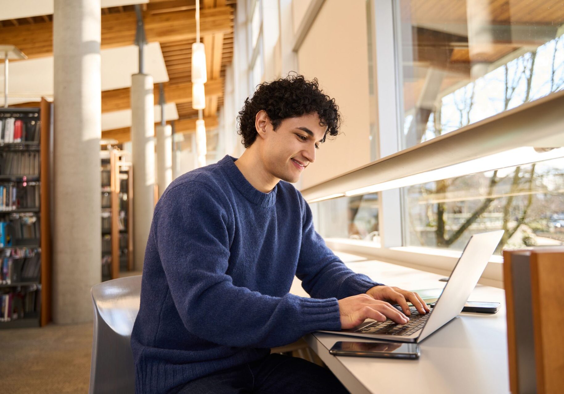 Smart Hispanic male student sitting at desk and using laptop preparing diploma project in the library campus