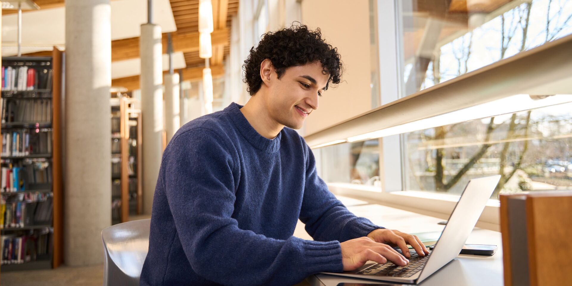 Smart Hispanic male student sitting at desk and using laptop preparing diploma project in the library campus