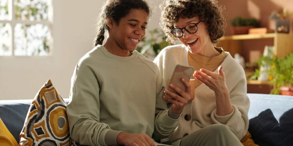 Happy mom spending time with her adopted daughter at home, they sitting on sofa and watching photos on mobile phone