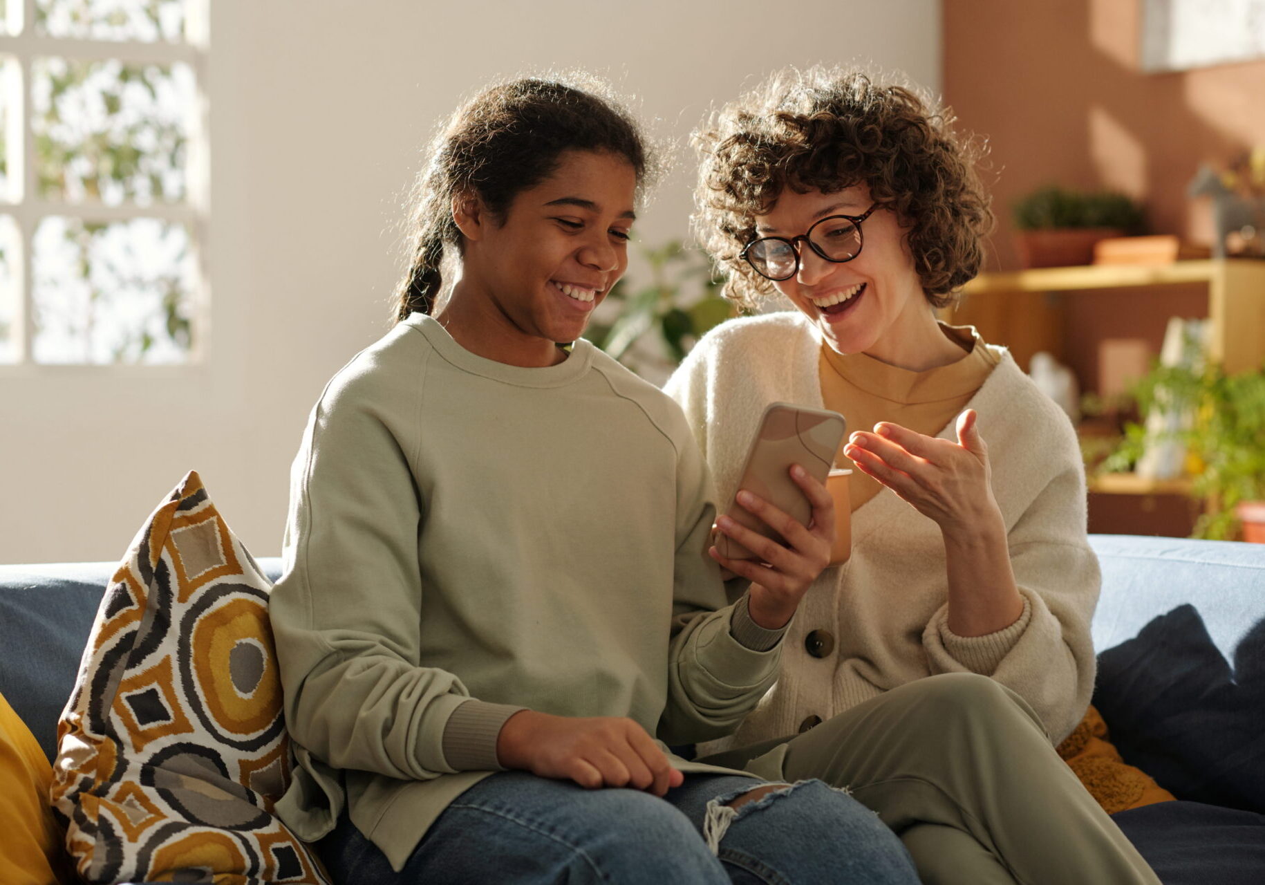 Happy mom spending time with her adopted daughter at home, they sitting on sofa and watching photos on mobile phone